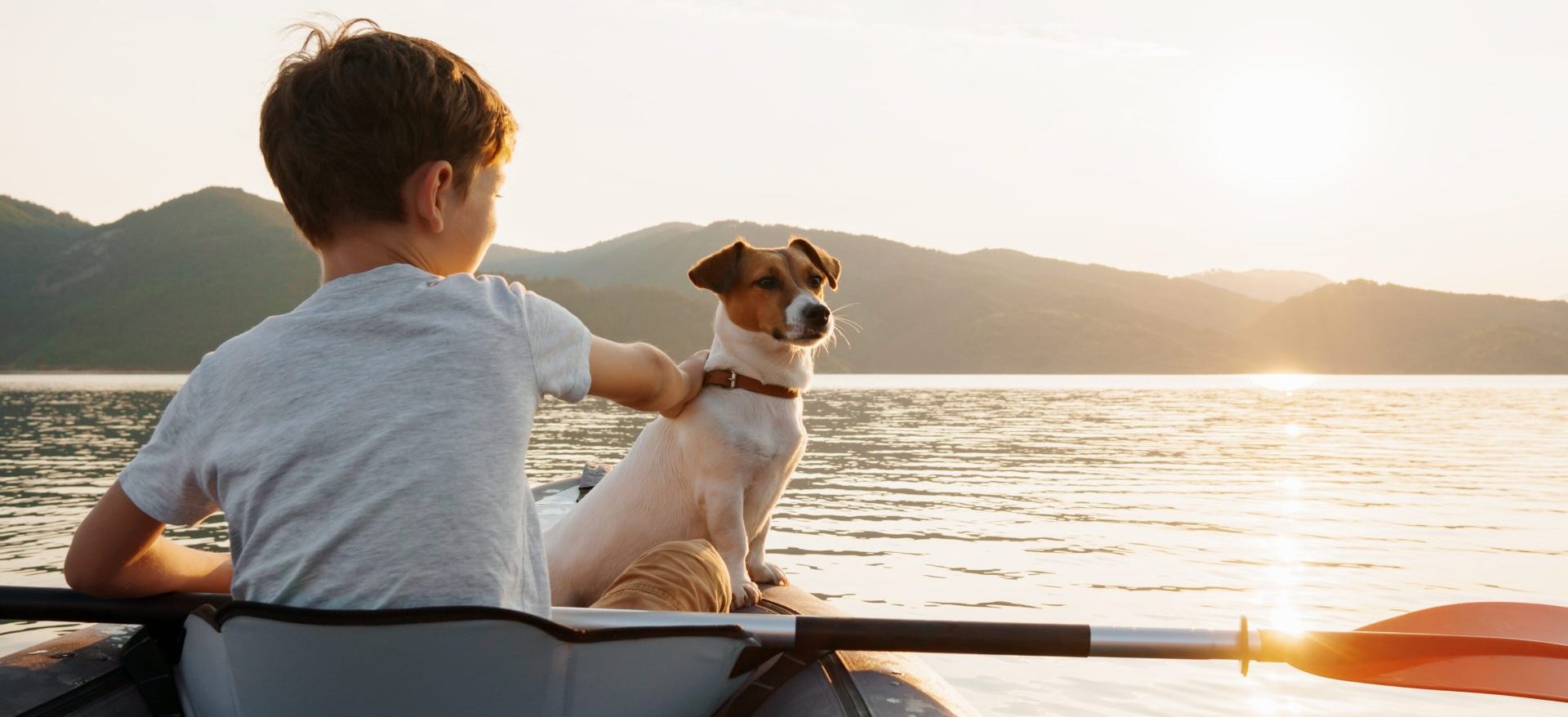 Happy boy with his dog Jack Russell Terrier paddling an inflatable kayak on the water mountain lake against the backdrop of beautiful orange sunset. Family sports vacation. Lens flare. Pet